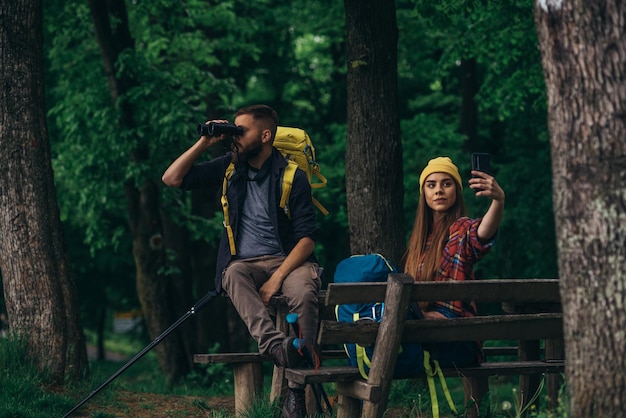 Randonneurs assis sur un banc dans la forêt tout en utilisant un smartphone et des jumelles