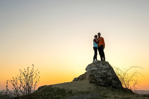 Randonneurs homme et femme debout sur une grosse pierre au coucher du soleil dans les montagnes Couple ensemble sur un haut rocher dans la nature du soir Tourisme voyage et concept de mode de vie sain