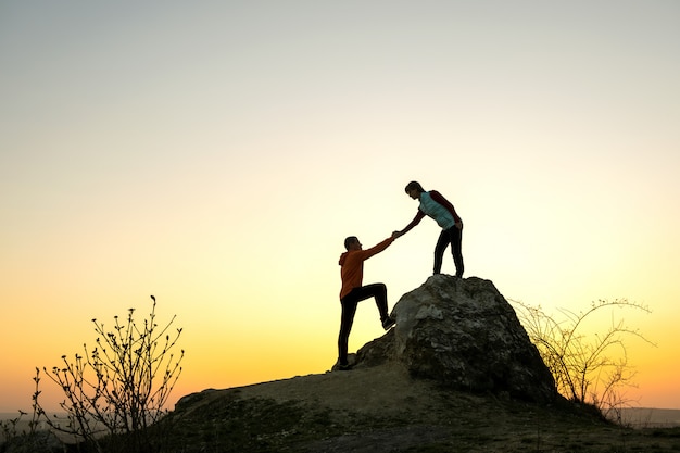 Randonneurs homme et femme s'aidant à gravir une grosse pierre au coucher du soleil dans les montagnes. Couple escalade sur un haut rocher dans la nature du soir. Concept de tourisme, de voyage et de mode de vie sain.