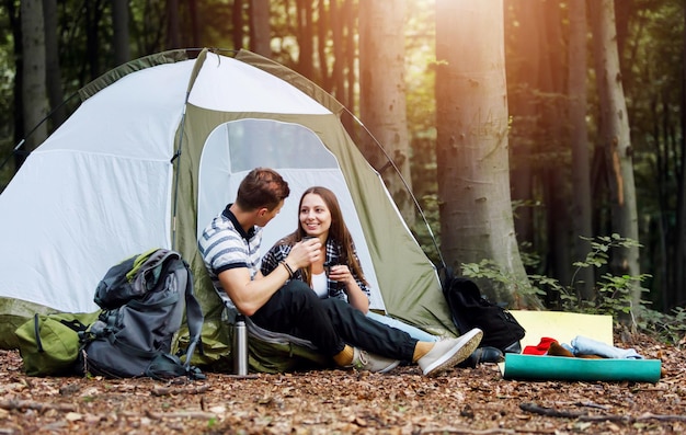 Randonneurs homme et femme se reposant devant la tente dans la forêt sous le ciel sur fond de forêt