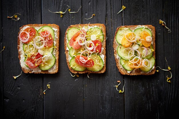 Rangée de trois toasts sains avec oignon concombre tomates cerises fromage feta émietté et pousses de radis sur une plaque noire Vue de dessus de table sur fond en bois noir