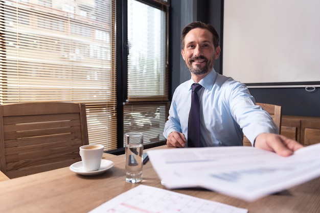 Rapports de travail. Beau gestionnaire responsable gai assis à la table et étendant la main tout en vous donnant les documents