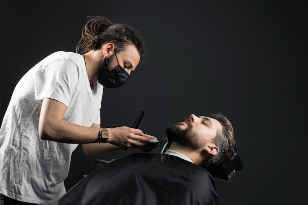 Rasage de la barbe dans le salon de coiffure. Barbier avec des dreadlocks dans un masque médical noir tailler un homme barbu en quarantaine coronavirus covid-19.