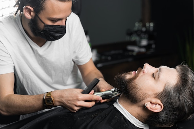 Rasage de la barbe dans le salon de coiffure. Barbier avec des dreadlocks dans un masque médical noir tailler un homme barbu en quarantaine coronavirus covid-19.