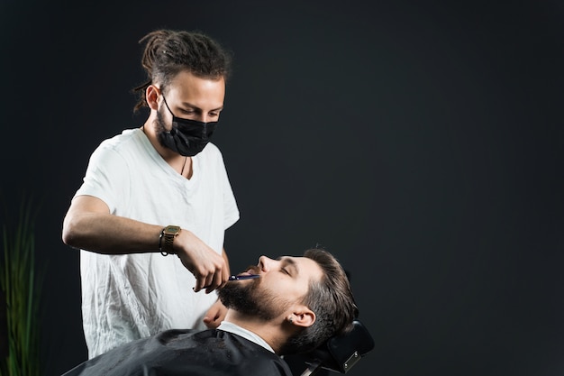 Rasage de la barbe dans le salon de coiffure. Barbier avec des dreadlocks dans un masque médical noir tailler un homme barbu en quarantaine coronavirus covid-19.