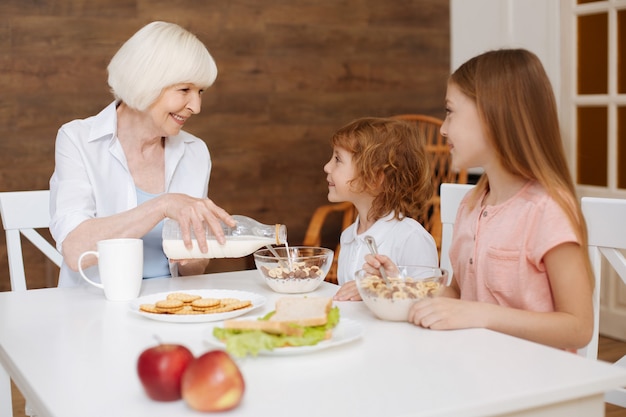 Rassemblement De Famille Positif Lumineux Actif à La Table Pour Prendre Le Petit-déjeuner Pendant Que Grand-mère Pore Du Lait Frais Dans Leurs Bols