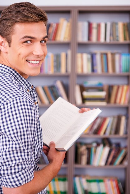 Rat de bibliothèque heureux. Vue arrière d'un jeune homme heureux tenant un livre et regardant par-dessus l'épaule en se tenant debout contre une étagère