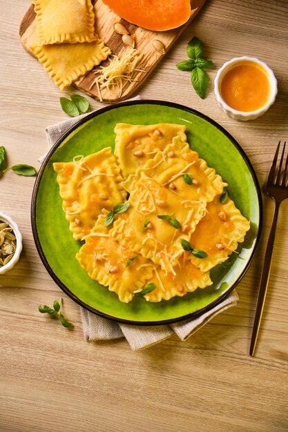 Des raviolis à la citrouille faits maison avec du fromage ricotta, du basilic, des pignons de pin et du fromage à pâte dure sur une plaque verte entourée d'ingrédients sur une table en bois. Concept de nourriture d'automne. Mise au point sélective.