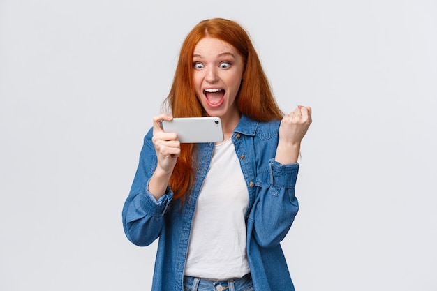 Réalisation, objectif et concept de réussite. Enthousiaste heureux mignon rousse adolescente pompe à poing triomphant, regardant l'écran du smartphone étonné et satisfait, remportant le défi du jeu