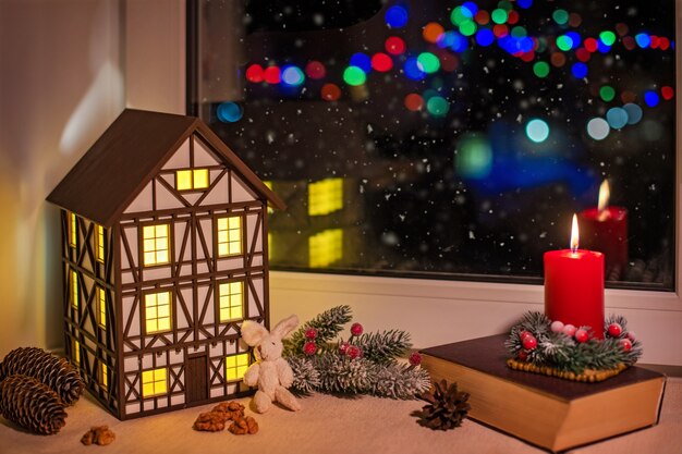 Sur le rebord de la fenêtre, il y a une veilleuse en forme de maison à colombages parmi les décorations de Noël, une bougie rouge brûle