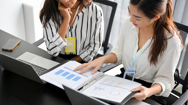 Recadrée de deux jeunes collègues travaillant sur des documents de données financières tout en utilisant un ordinateur portable.