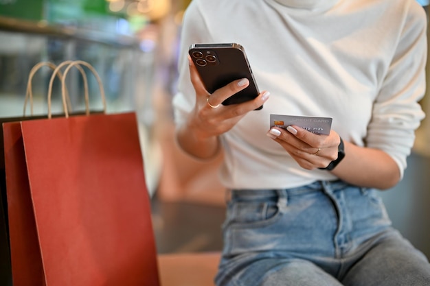 Recadrée une femme tenant une carte de crédit et utilisant son smartphone lors de ses achats au centre commercial