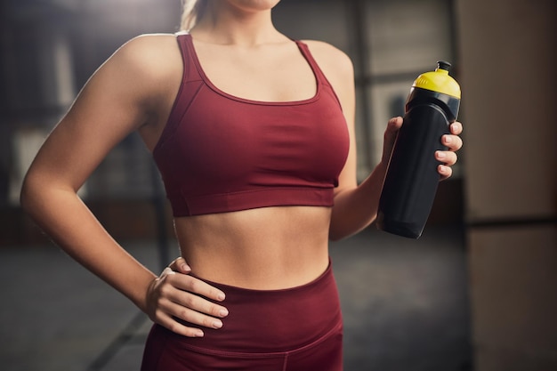 Recadrer une athlète féminine mince et méconnaissable en tenue de sport avec la main sur la hanche et une bouteille d'eau dans la salle de sport