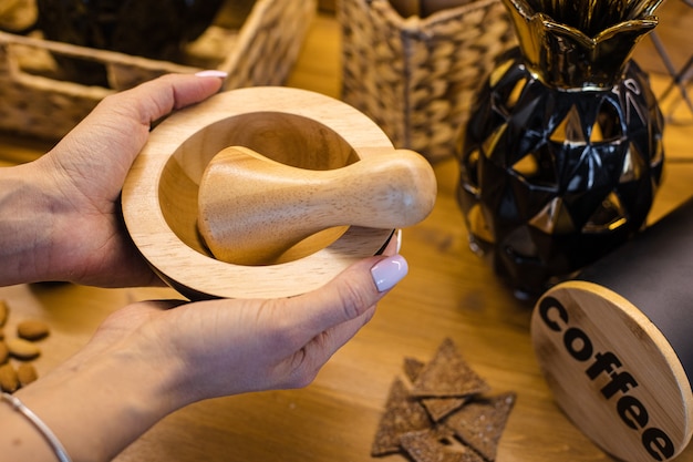 Recadrer la photo d'une femme méconnaissable tenant un mortier et un pilon en bois pour moudre le café. Elle le tient au-dessus de la table en bois avec un récipient, dit du café et des craquelins sur la table.