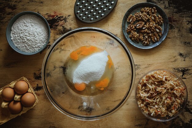 Recette de gâteau aux pommes et aux carottes et aux noix. Oeufs et sucre dans un bol. Ingrédients sur la table en bois. Farine, œufs, râpe.