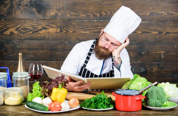 Photo recette pour cuisiner des aliments sains chef expérimenté cuisinant un excellent plat cette recette est tout simplement parfaite recette végétarienne arts culinaires homme barbu hipster lire la recette du livre près de la table légumes frais