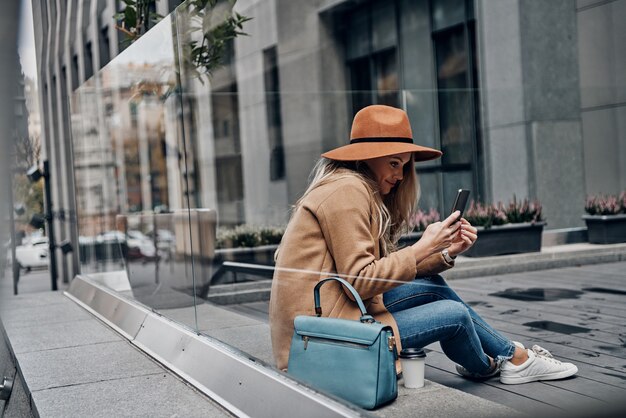 Recevoir de beaux messages. À travers la vitrine d'une jolie jeune femme en chapeau et manteau utilisant son téléphone intelligent tout en passant du temps sans soucis