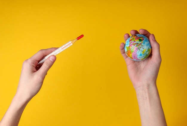 Réchauffement climatique. Mains féminines tenant un thermomètre et un globe sur fond jaune. Vue de dessus