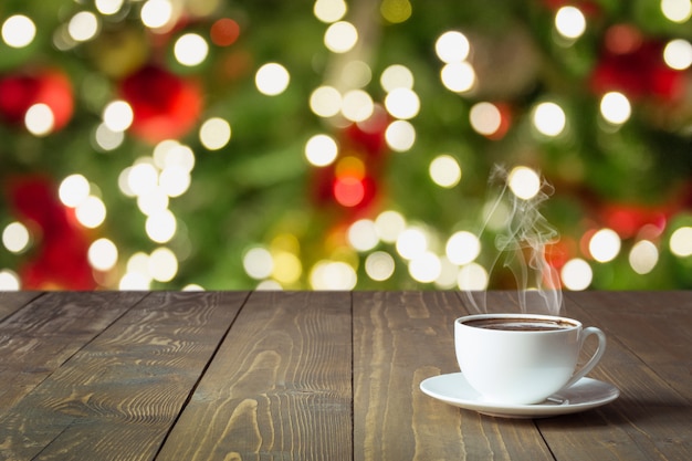 Réchauffer Une Tasse De Café Noir Sur Une Table En Bois. Arbre De Noël Flou Comme Toile De Fond. Période De Noël.