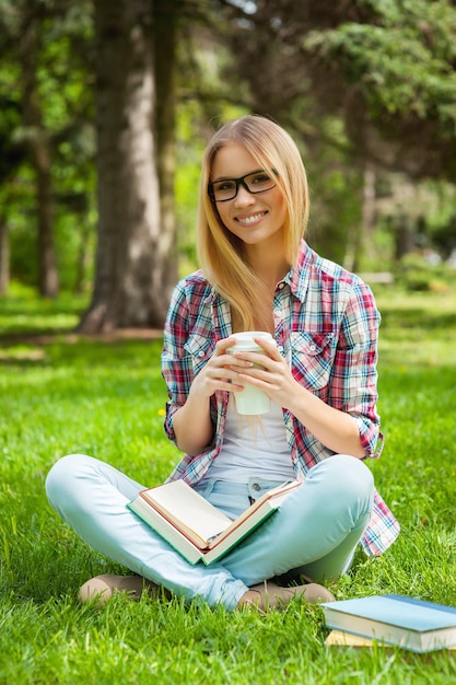 Recherche d'inspiration à l'extérieur. Belle jeune étudiante ajustant ses lunettes et souriant alors qu'elle était assise dans un parc avec des livres autour d'elle