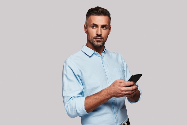 A la recherche d'une réponse. Beau jeune homme en tenue décontractée intelligente à l'aide de son téléphone intelligent et souriant en se tenant debout sur fond gris