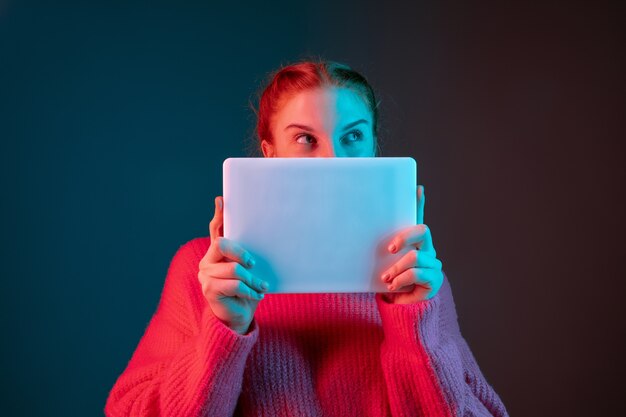 À la recherche sur tablette. Portrait de femme caucasienne isolé sur fond de studio dégradé à la lumière du néon. Beau modèle féminin aux cheveux rouges. Concept d'émotions humaines, expression faciale, annonce.