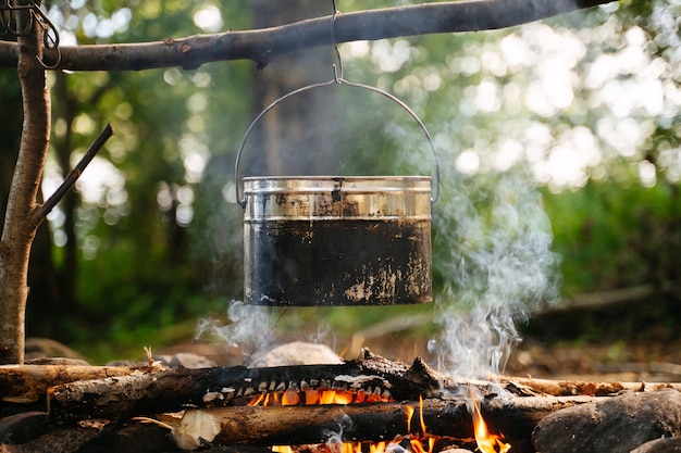 Un récipient de cuisson extérieur en aluminium est suspendu à un bâton en bois au-dessus du feu