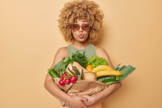 Récolte de printemps Une jeune femme aux cheveux bouclés garde les lèvres pliées embrasse le bouquet avec des légumes et des fruits frais cueillis pour faire un plat végétarien porte des lunettes de soleil isolées sur fond marron