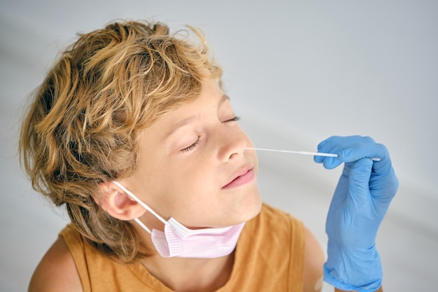 Récolter un médecin méconnaissable dans un gant stérile avec un écouvillon prenant le test COVID 19 du nez d'un enfant aux yeux fermés à l'hôpital