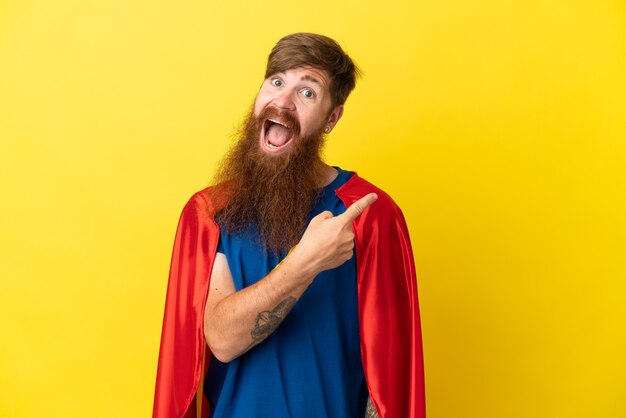 Redhead Super Hero man isolé sur fond jaune surpris et pointant du côté