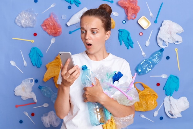 Réduire la pollution par les ordures Nettoyer la planète Choqué femme surprise portant une chemise blanche isolée sur un mur bleu avec des ordures autour en regardant l'écran du téléphone portable avec la bouche ouverte