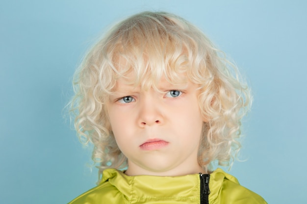 Réfléchi. Bouchent le portrait du beau petit garçon caucasien isolé sur le mur bleu. Modèle masculin blond bouclé. Concept d'expression faciale, émotions humaines, enfance,