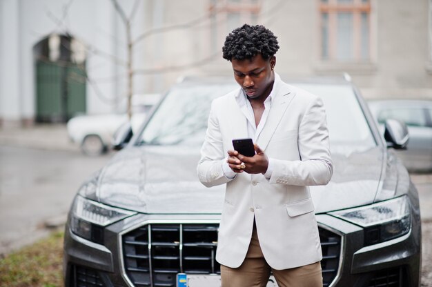 Réfléchi jeune beau gentleman afro-américain en tenue de soirée. Homme modèle élégant noir en veste blanche avec téléphone portable à mains contre voiture d'affaires.