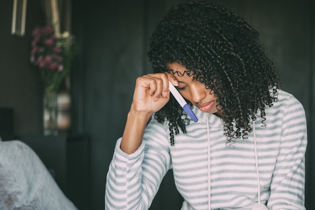 Réfléchie femme noire inquiète et triste avec test de grossesse au lit