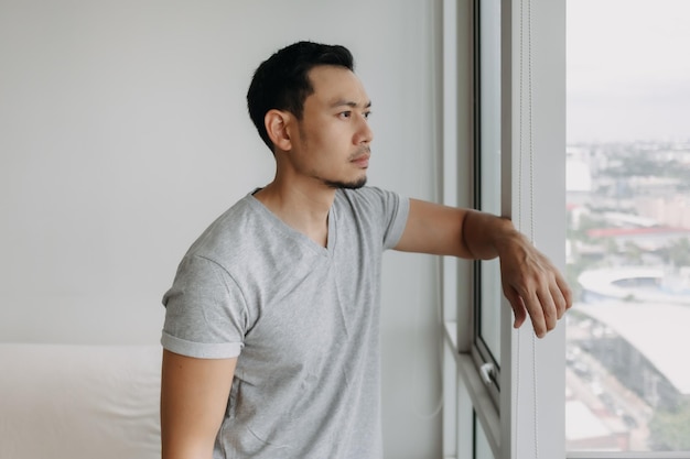 Réfléchissant sérieusement, un homme asiatique solitaire s'appuie sur la fenêtre de son appartement.