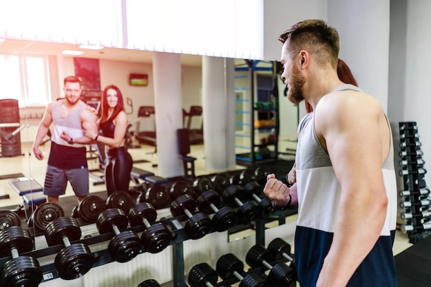 Reflet dans le miroir d'un couple sportif sur fond d'haltères noirs Belle femme et un homme regardant le miroir sur leurs corps musclés dans la salle de sport