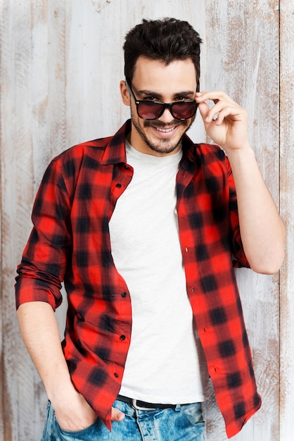 Regard séduisant. Beau jeune homme ajustant ses lunettes de soleil et regardant la caméra tout en se penchant sur le mur en bois