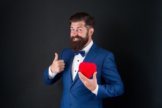 En regardant bien. Célébration des vacances. notion d'amour. Journée mondiale du cœur. hipster barbu brutal en costume formel. homme d'affaires en noeud papillon. Soins de santé. Décoration de la Saint-Valentin. homme de smoking avec coeur rouge.