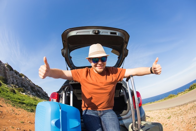 En regardant la caméra, un homme joyeux partant pour le week-end en voiture avec des bagages.