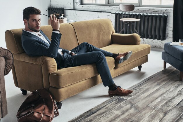 En regardant dans ton cœur. Beau jeune homme en costume complet tenant des lunettes et regardant la caméra alors qu'il était assis sur le canapé