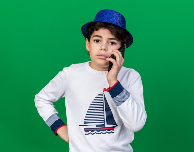En regardant devant un petit garçon portant un chapeau de fête bleu parle au téléphone en mettant la main sur la hanche isolée sur un mur vert
