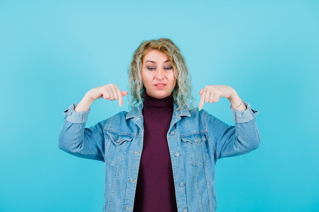 Regardant vers le bas, la femme blonde pointe vers le bas avec les index sur fond bleu