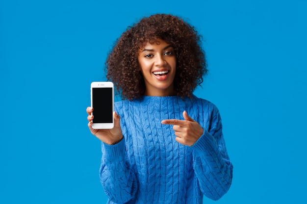 Regarde ça. Heureuse femme afro-américaine charismatique avec coupe de cheveux afro, tenant un smartphone, montrant un écran mobile, pointant l'affichage comme application de promotion, application de magasinage ou jeu