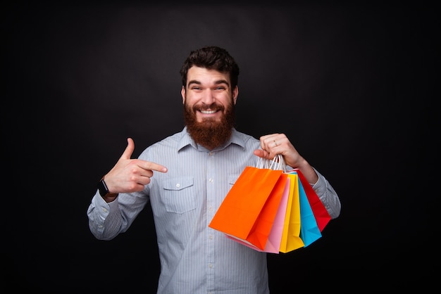 Regarde, j'ai des sacs de courses ! Un jeune homme barbu joyeux tient cinq sacs à provisions colorés en papier et les pointe du doigt.