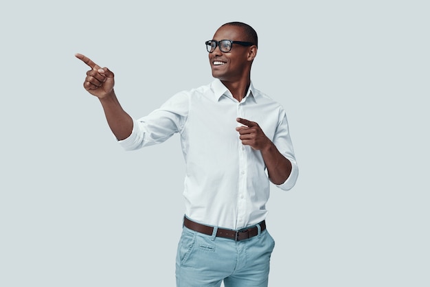 Regarde là-bas! Beau jeune homme africain pointant l'espace de copie et souriant en se tenant debout sur fond gris