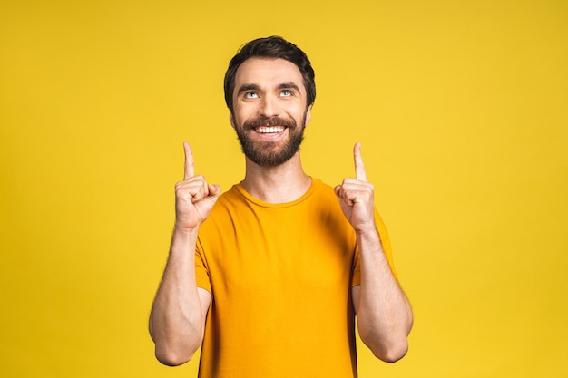 Regarde là-bas! Heureux jeune bel homme barbu décontracté pointant vers le haut et souriant tout en se tenant isolé sur fond jaune.