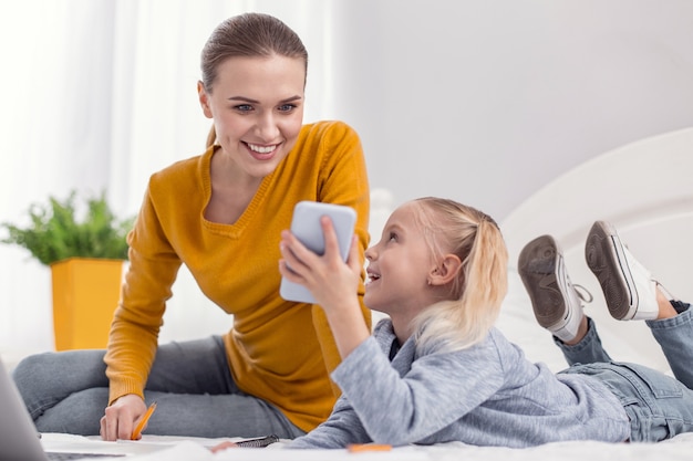 Regarde maman. Joyeuse mère énergique regardant l'écran tout en fille mignonne montrant son téléphone et riant