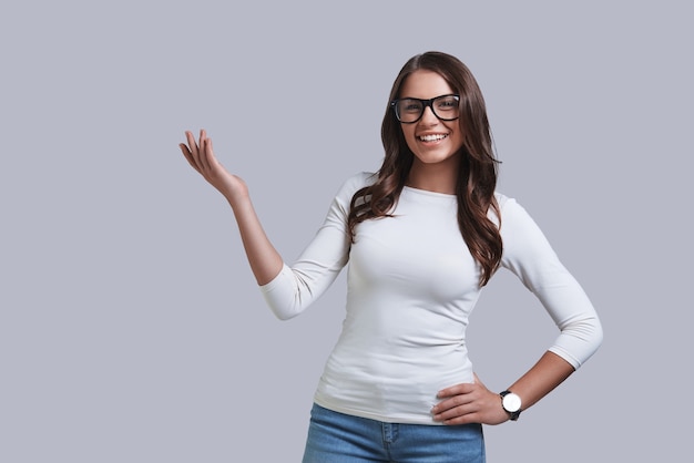 Regarde par ici ! Belle jeune femme pointant du doigt et souriant en se tenant debout sur fond gris