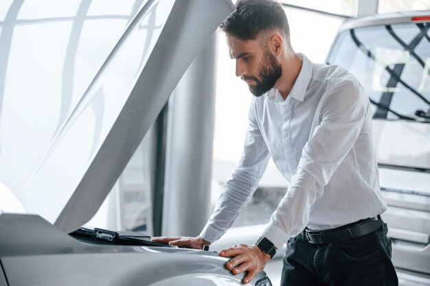 Regarde sous le capot de l'automobile Jeune homme en vêtements blancs est dans le concessionnaire automobile