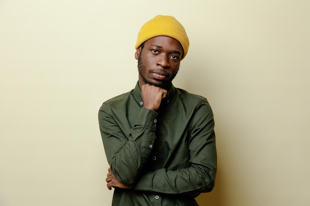 Regarder la caméra en mettant la main sur le menton jeune homme afro-américain au chapeau portant une chemise verte isolée sur fond blanc